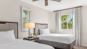 Ein Hotelzimmer mit zwei Betten, einem Deckenventilator, einem Fenster mit Vorhängen und einem Nachttisch mit Lampe und Blumenvase.