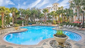 Ein Hotelpool mit Liegestühlen, Palmen und einem mehrstöckigen Gebäude im Hintergrund.
