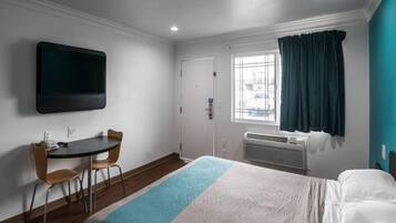 Habitación de hotel con cama, escritorio con silla, televisor de pantalla plana montado en la pared, ventana con cortinas y unidad de aire acondicionado.
