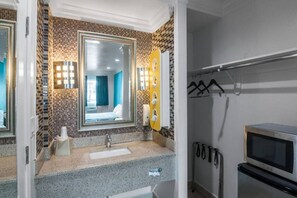 A bathroom with a tiled backsplash, a sink, a mirror, a towel dispenser, and a shower area.