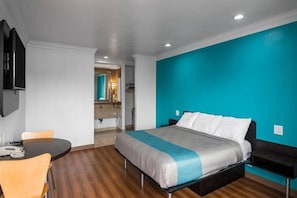 A modern hotel room with a bed, a desk, and a chair. There is a bathroom visible in the background.