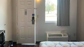A hotel room with a bed, a door, a window with curtains, a radiator, and a wall-mounted air conditioning unit.