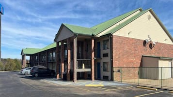 A two-story building with a green roof, a brick facade, and a covered entrance area. There is a parking lot with marked spaces and a fence surrounding the property.