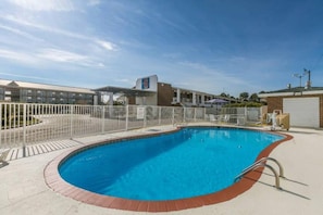 Una piscina con cerco blanco, borde de ladrillo y cielo azul despejado al fondo.