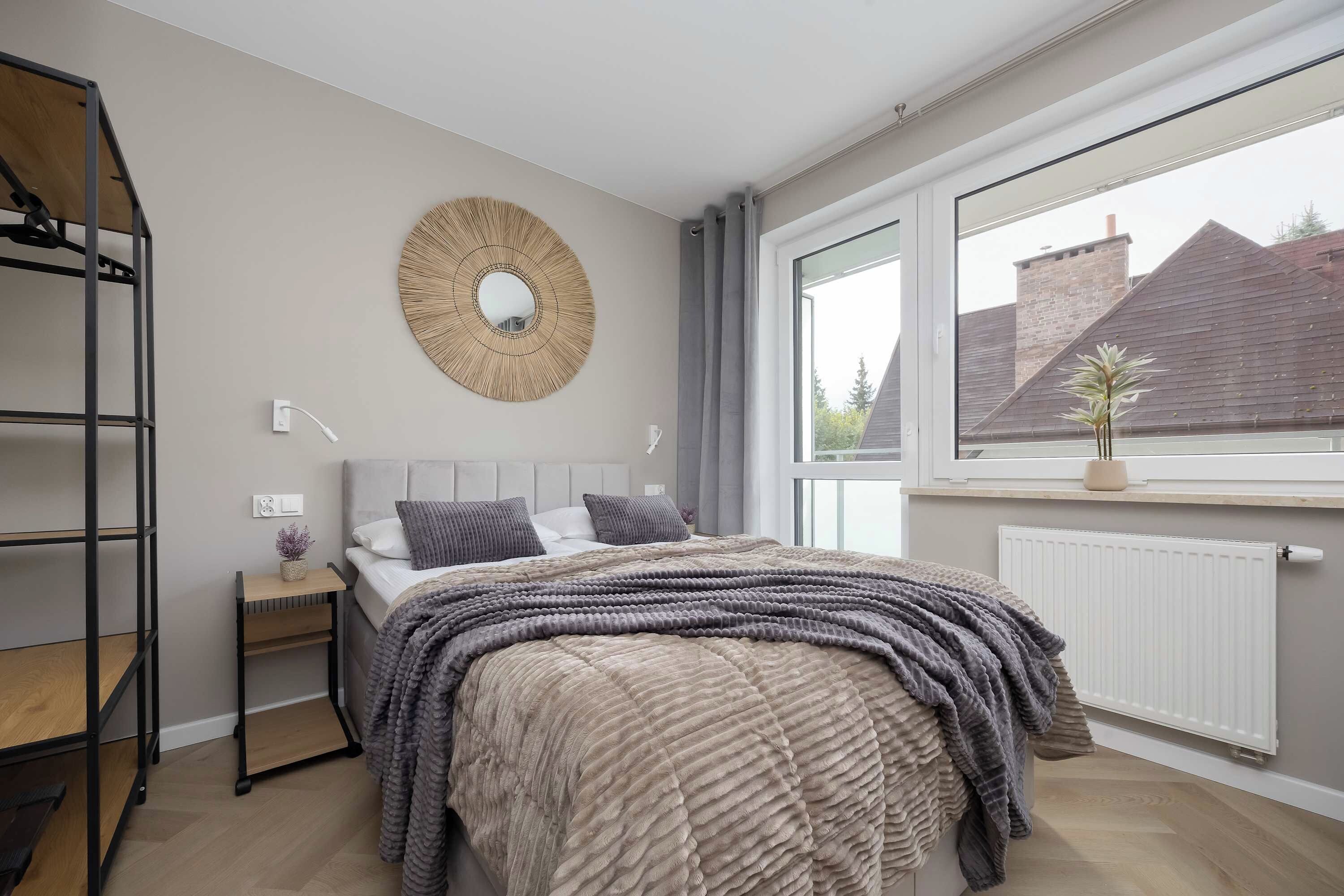 Une chambre à coucher comprenant un lit, une table de chevet, un miroir fixé au mur et une fenêtre donnant sur des immeubles.