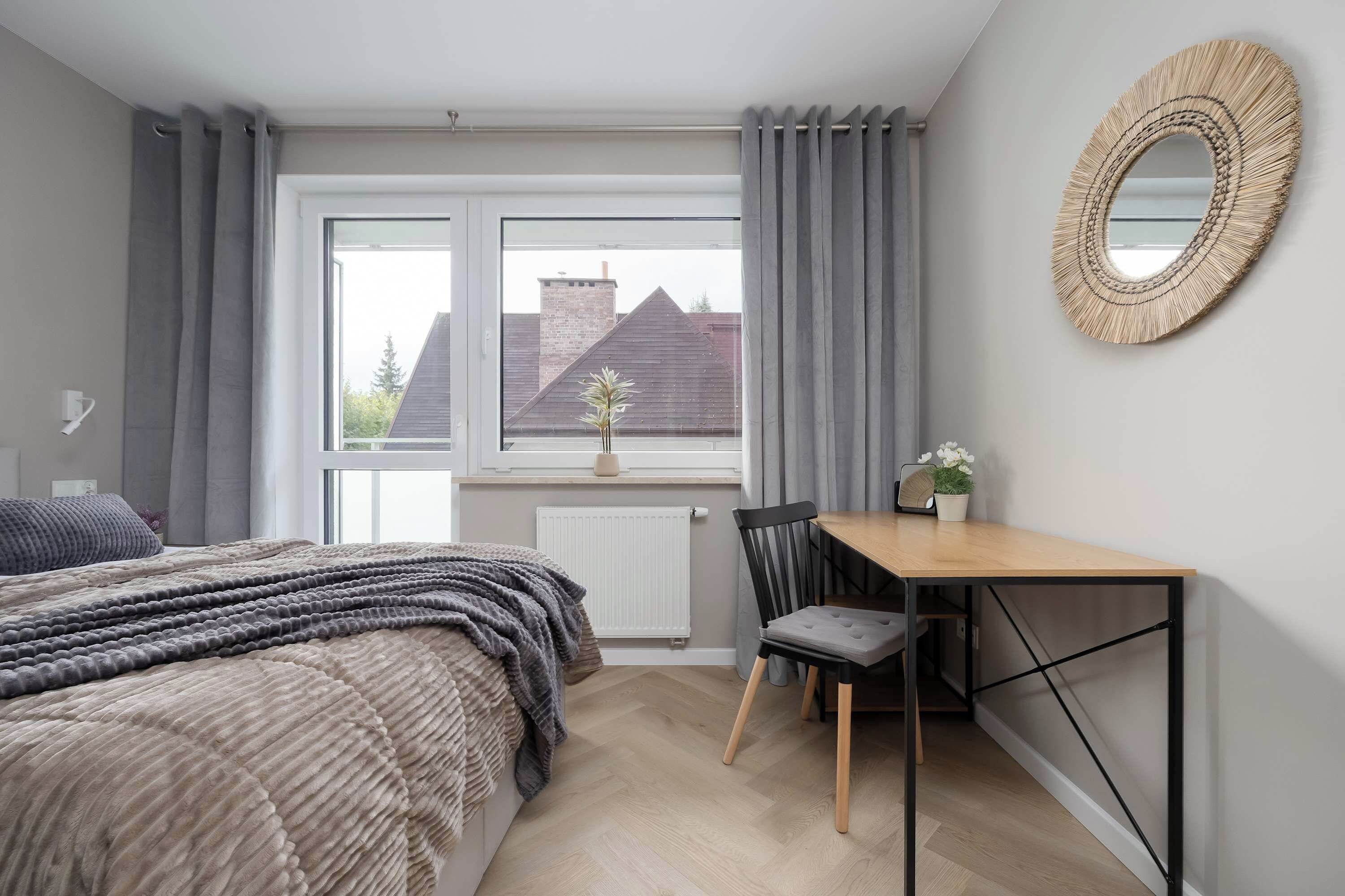 A bedroom with a bed, a desk, and a chair. There is a window with curtains, a radiator, and a view of a building outside.