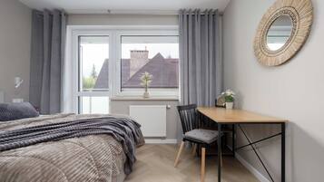 A bedroom with a bed, a desk, and a chair. There is a window with curtains, a radiator, and a view of a building outside.