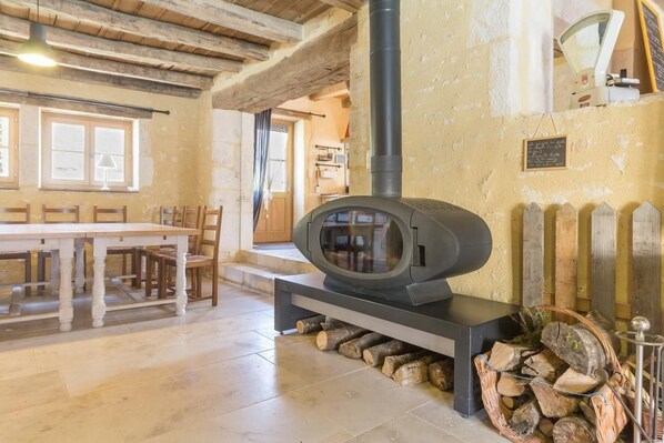 A rustic interior with a stone wall, a wooden table, chairs, a fireplace with logs, and a chalkboard with writing.