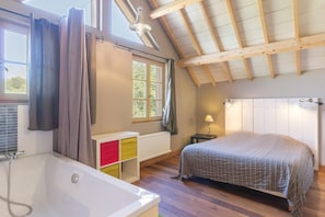 A bedroom with a sloped ceiling, a large window, a bed, a nightstand, a lamp, a bathroom with a bathtub, and a colorful cabinet.