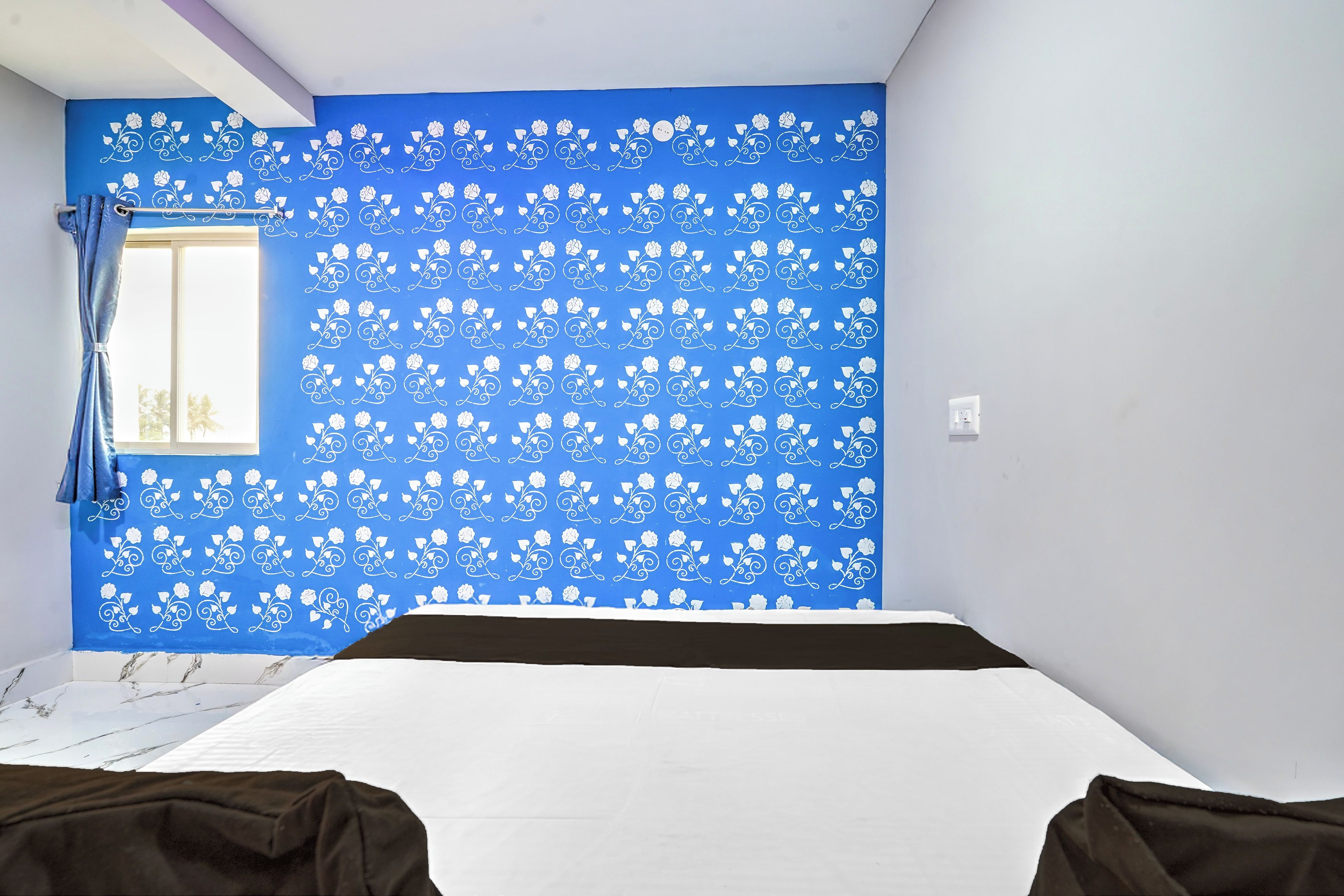 A room with a blue wall featuring a white floral pattern, a window with curtains, and a bed with a white mattress and brown headboard.