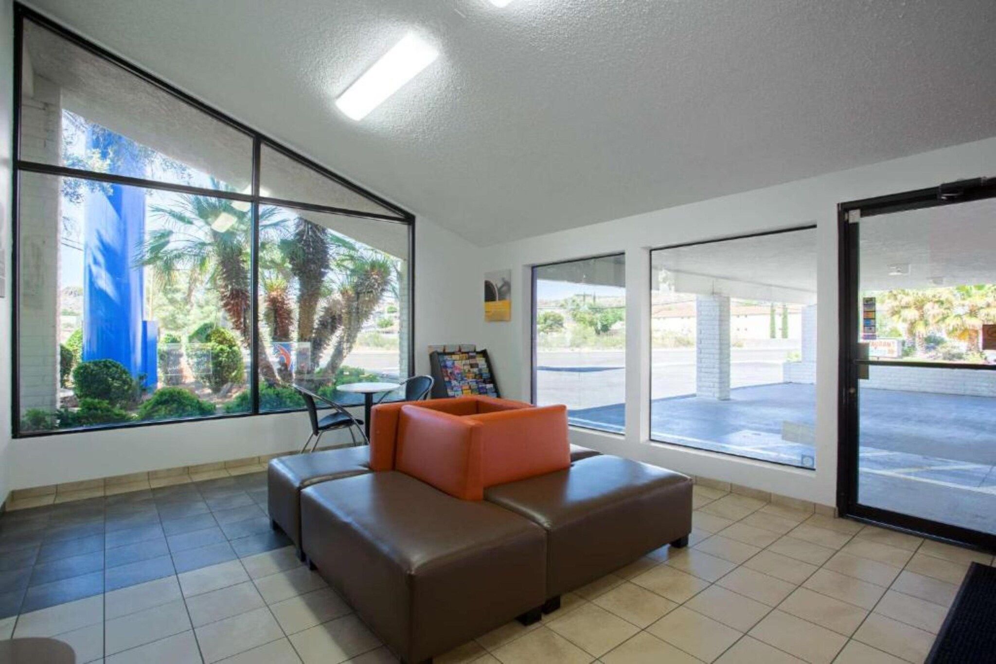 A lobby with a brown sofa, a table, and a chair. There are large windows with a view of palm trees and a building.
