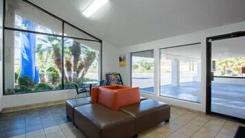 A lobby with a brown sofa, a table, and a chair. There are large windows with a view of palm trees and a building.