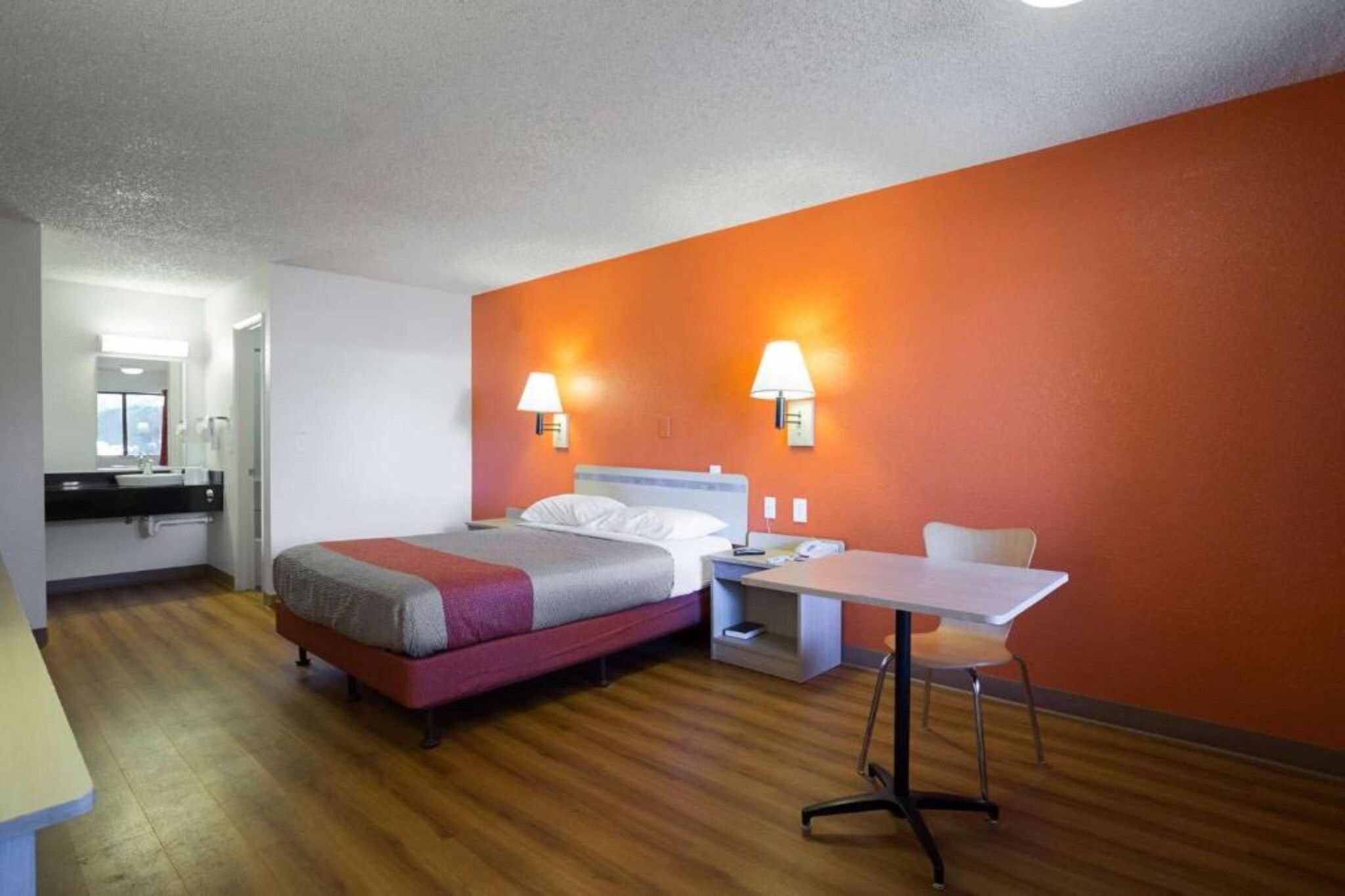 A hotel room with a bed, bedside table, chair, and a small table with a lamp.