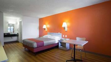A hotel room with a bed, bedside table, chair, and a small table with a lamp.