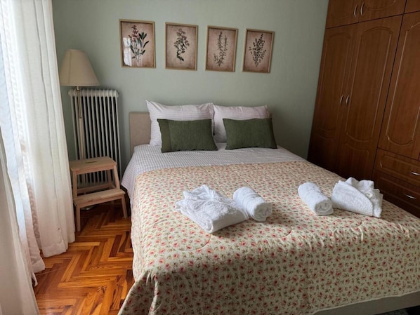 Ein ordentlich bezogenes Bett mit floralem Bettzeug, ein Nachttisch aus Holz, eine Heizung und ein Kleiderschrank.