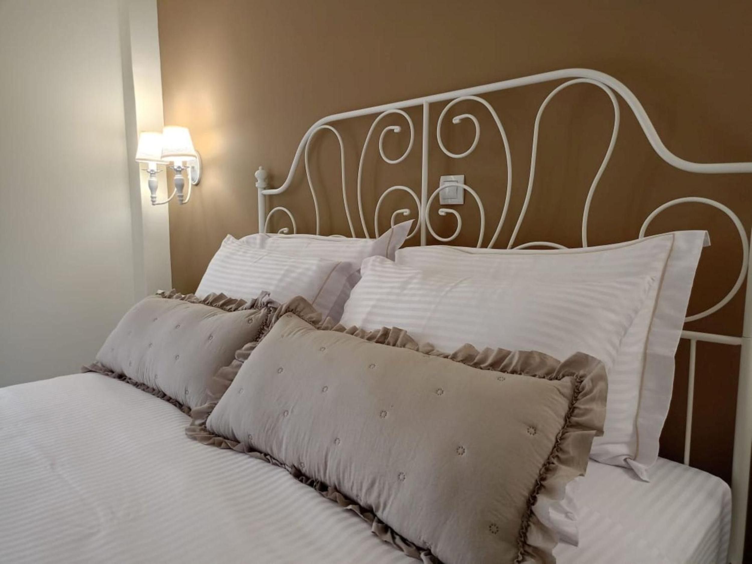 Een net opgemaakt bed met een wit metalen hoofdbord, twee kussens en een wandlamp.