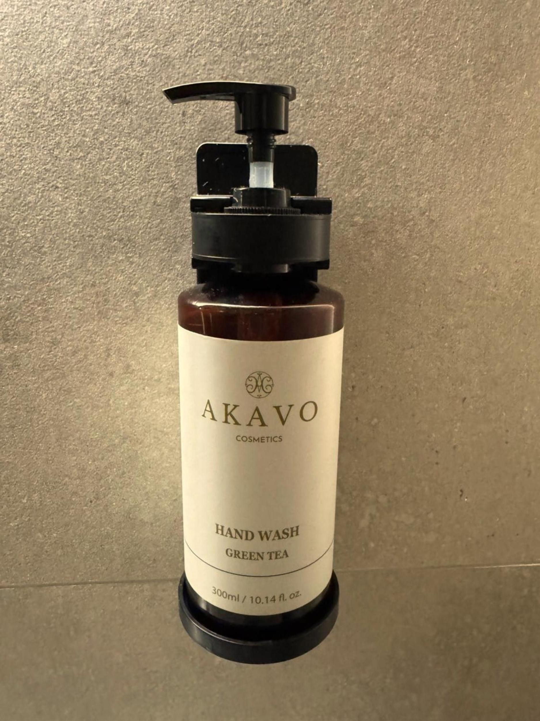 A hand sanitizer bottle labeled 'AKAVO COSMETICS HAND WASH GREEN TEA' with a pump dispenser, placed on a surface against a textured wall.