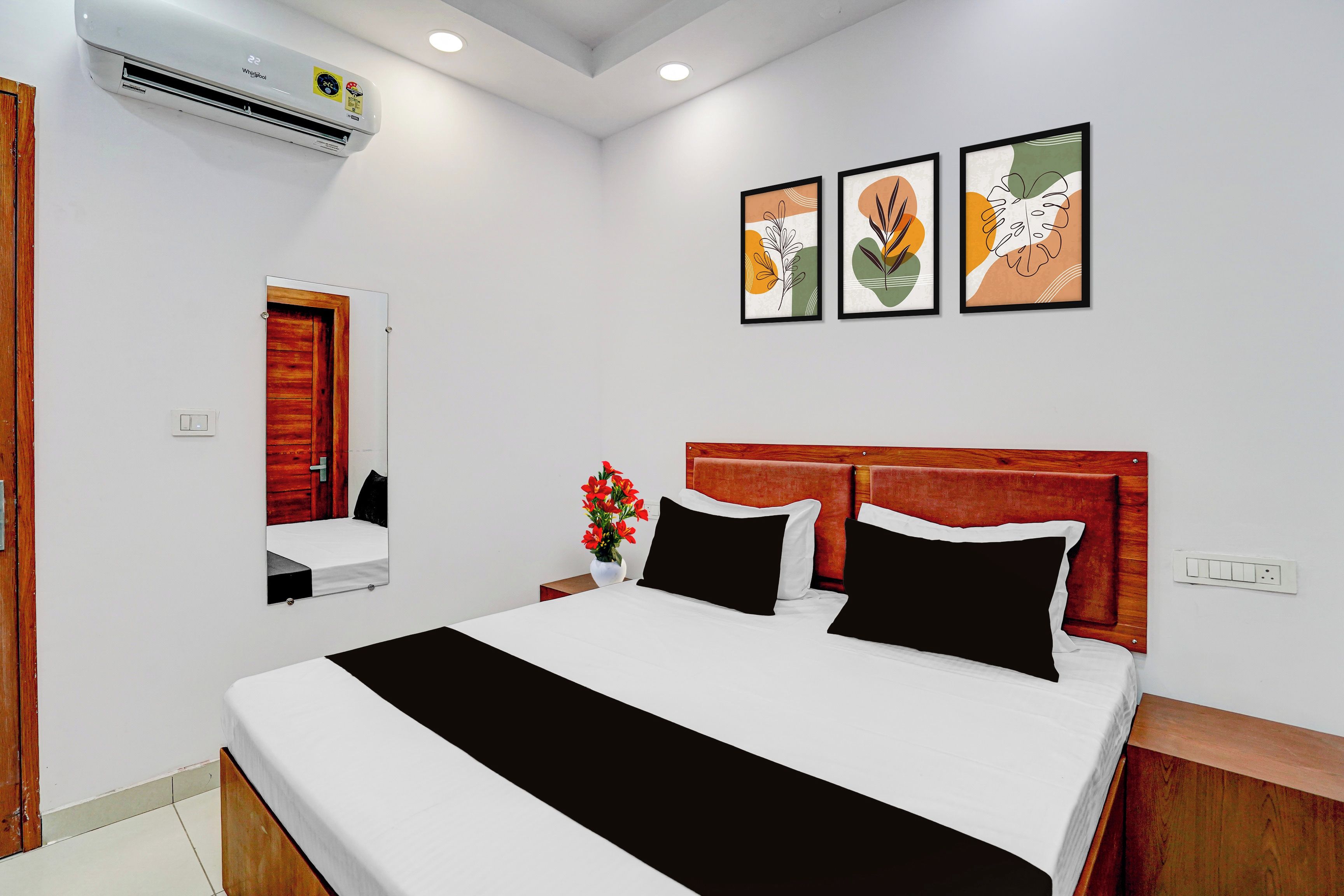 A hotel room with a bed, a bedside table with a vase of flowers, a wall-mounted air conditioner, and three framed artworks above the bed.