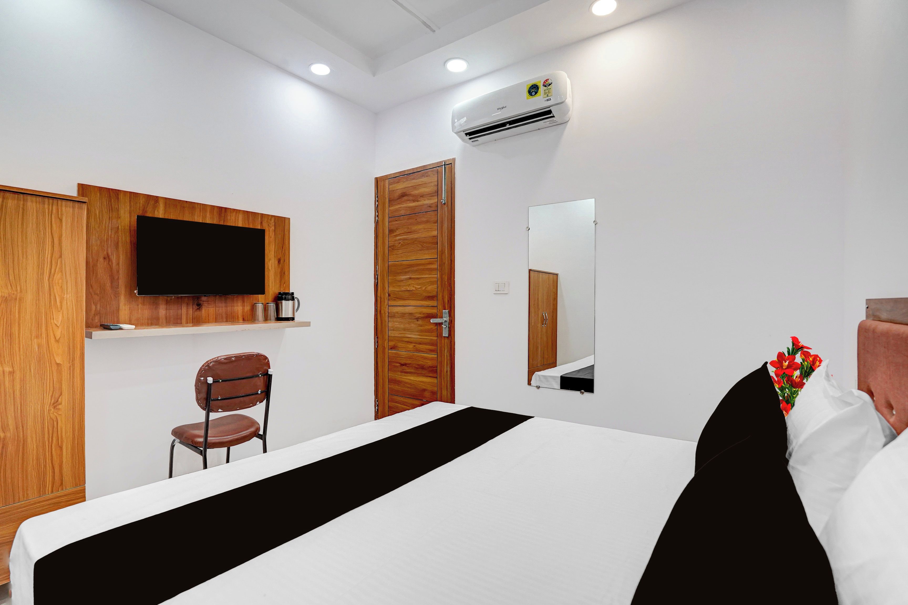 A hotel room with a bed, a wooden wardrobe, a television mounted on the wall, a mirror, and a small chair.