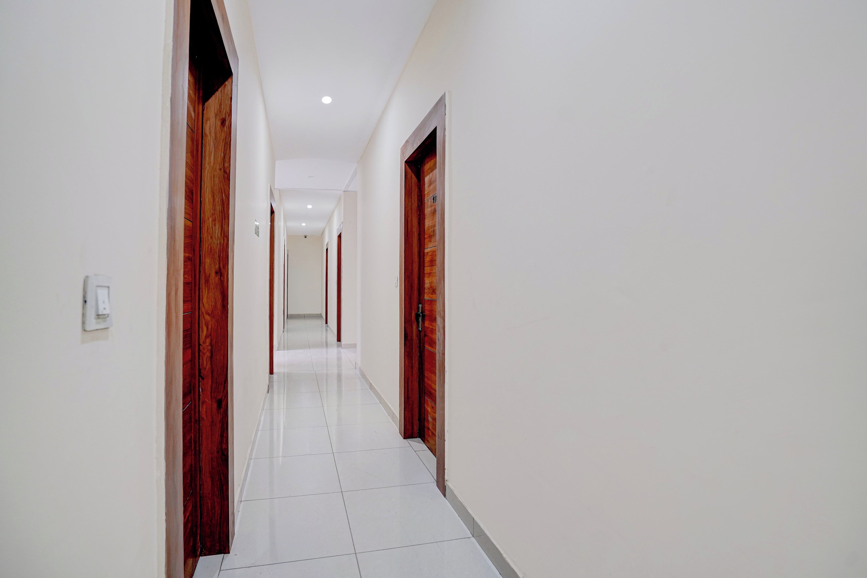 A corridor with white tiled flooring, wooden doors, and recessed ceiling lighting.