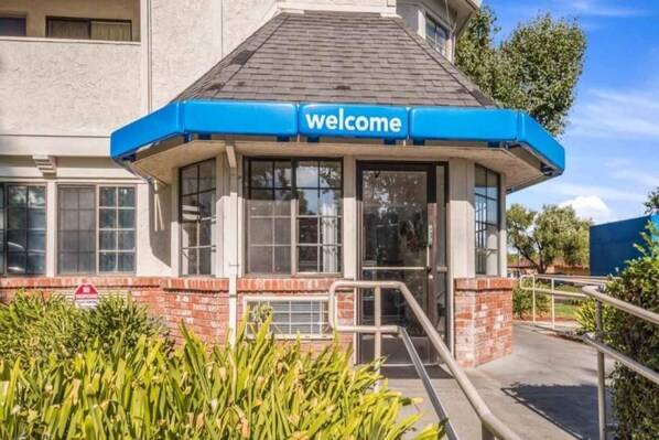 Une entrée d'immeuble avec une pancarte bleue portant l'inscription « Bienvenue », une façade en briques et une porte en verre.