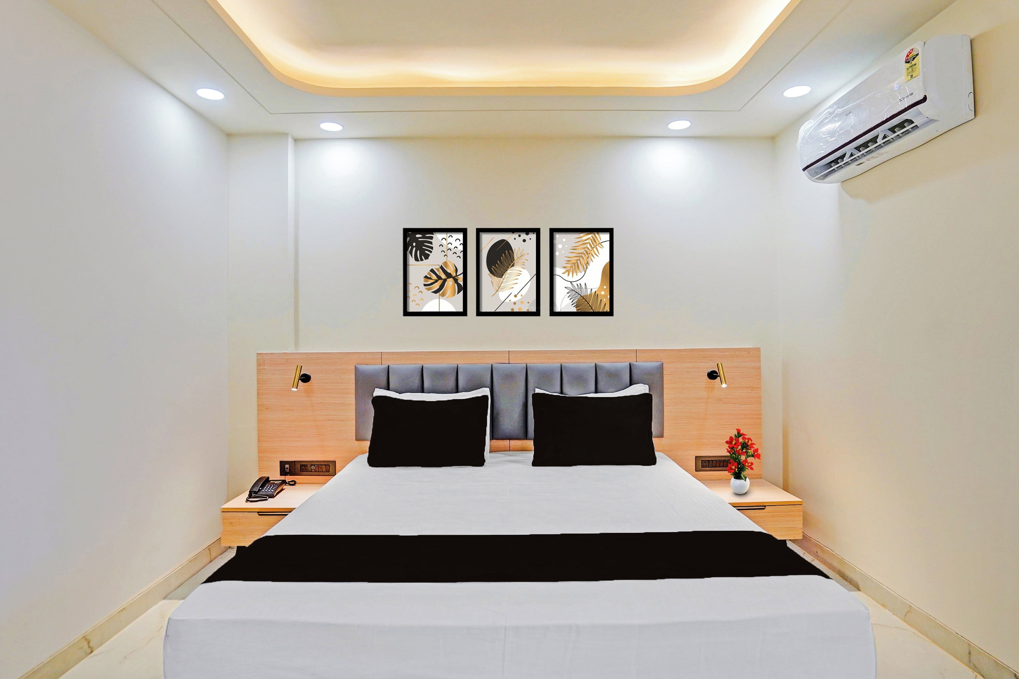 A modern hotel room with a large bed, two bedside tables, a nightstand, a wall-mounted air conditioner, and three framed artworks above the bed.