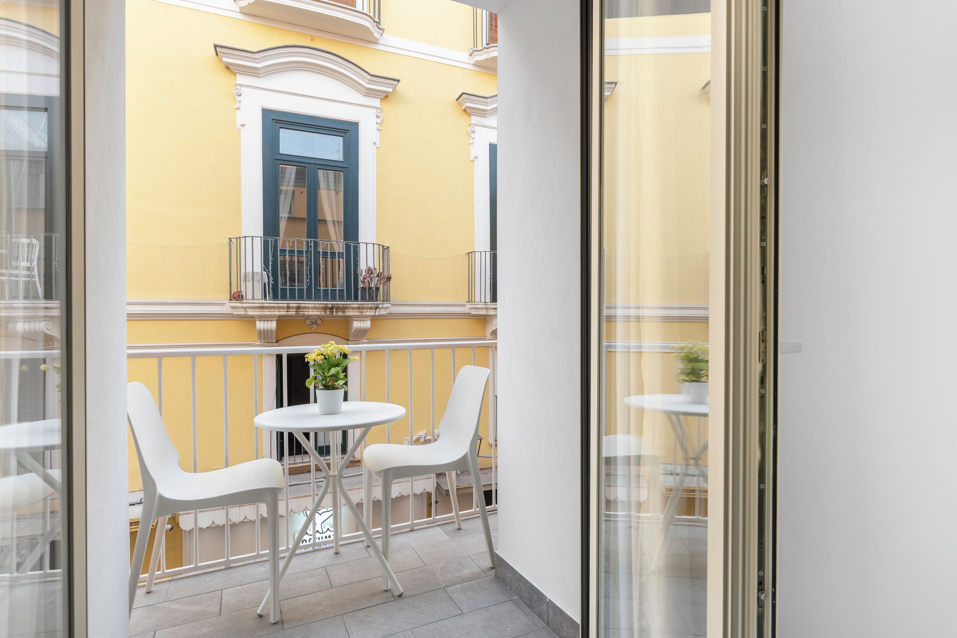 A balcony with white chairs and a small table, a potted plant, and a window with a blue frame.