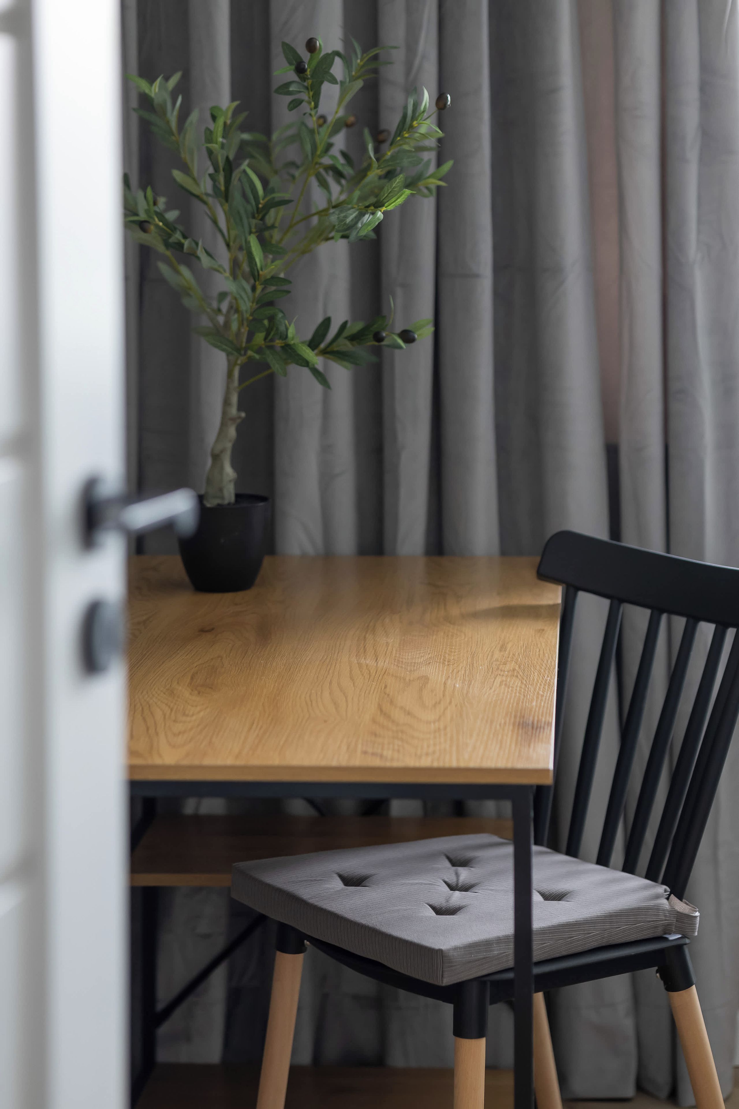 A wooden dining table with a potted plant, a black chair with a cushioned seat, and a curtain in the background.
