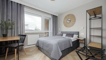 A modern bedroom with a bed, a desk, a chair, a radiator, a window with curtains, and a wall-mounted shelf.