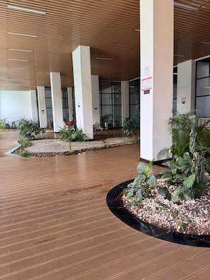Edificio moderno con piso de madera y columnas, rodeado de plantas en macetas y un sendero.
