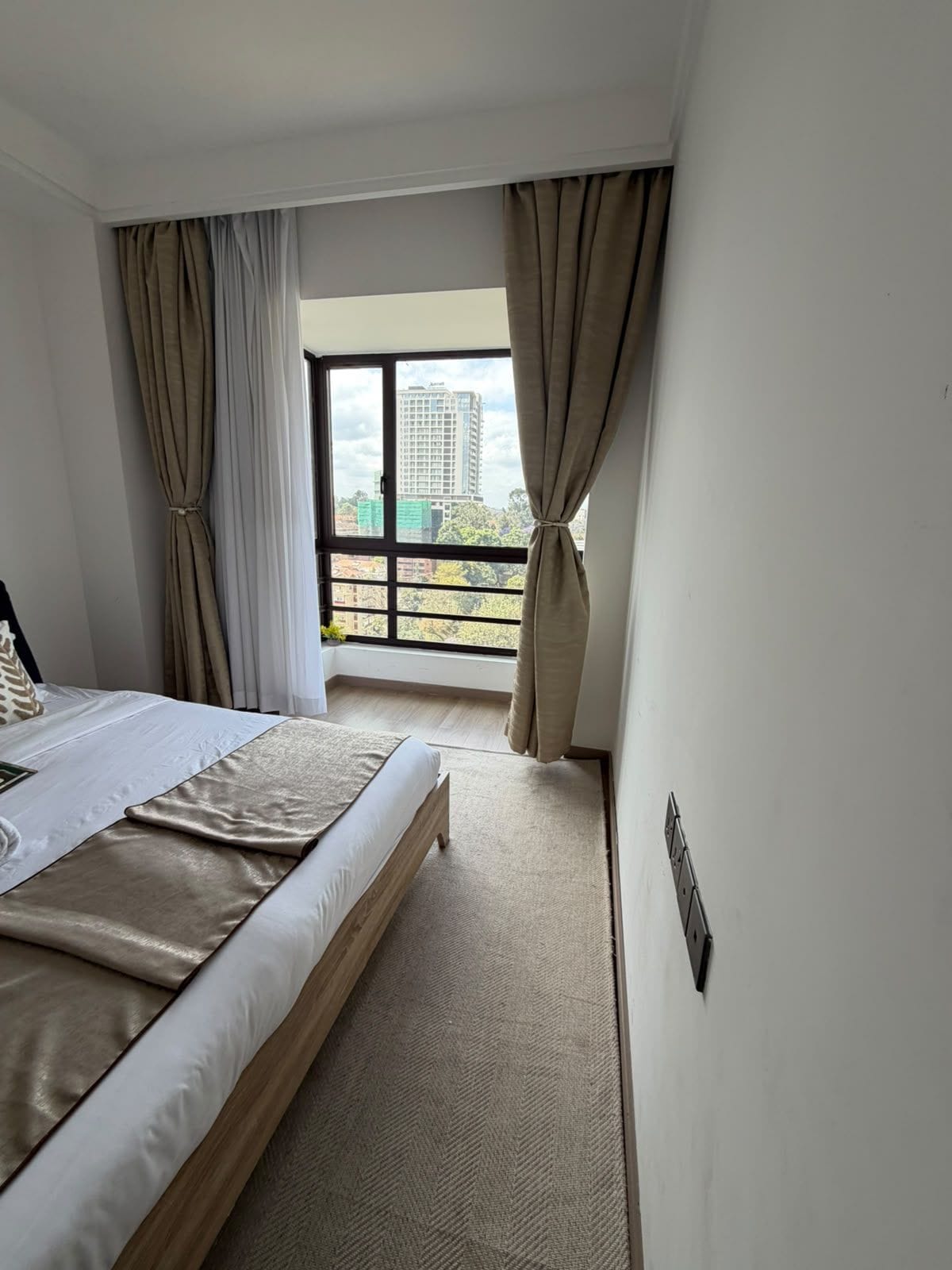 A hotel room with a bed, curtains, a window with a city view, and a carpeted floor.