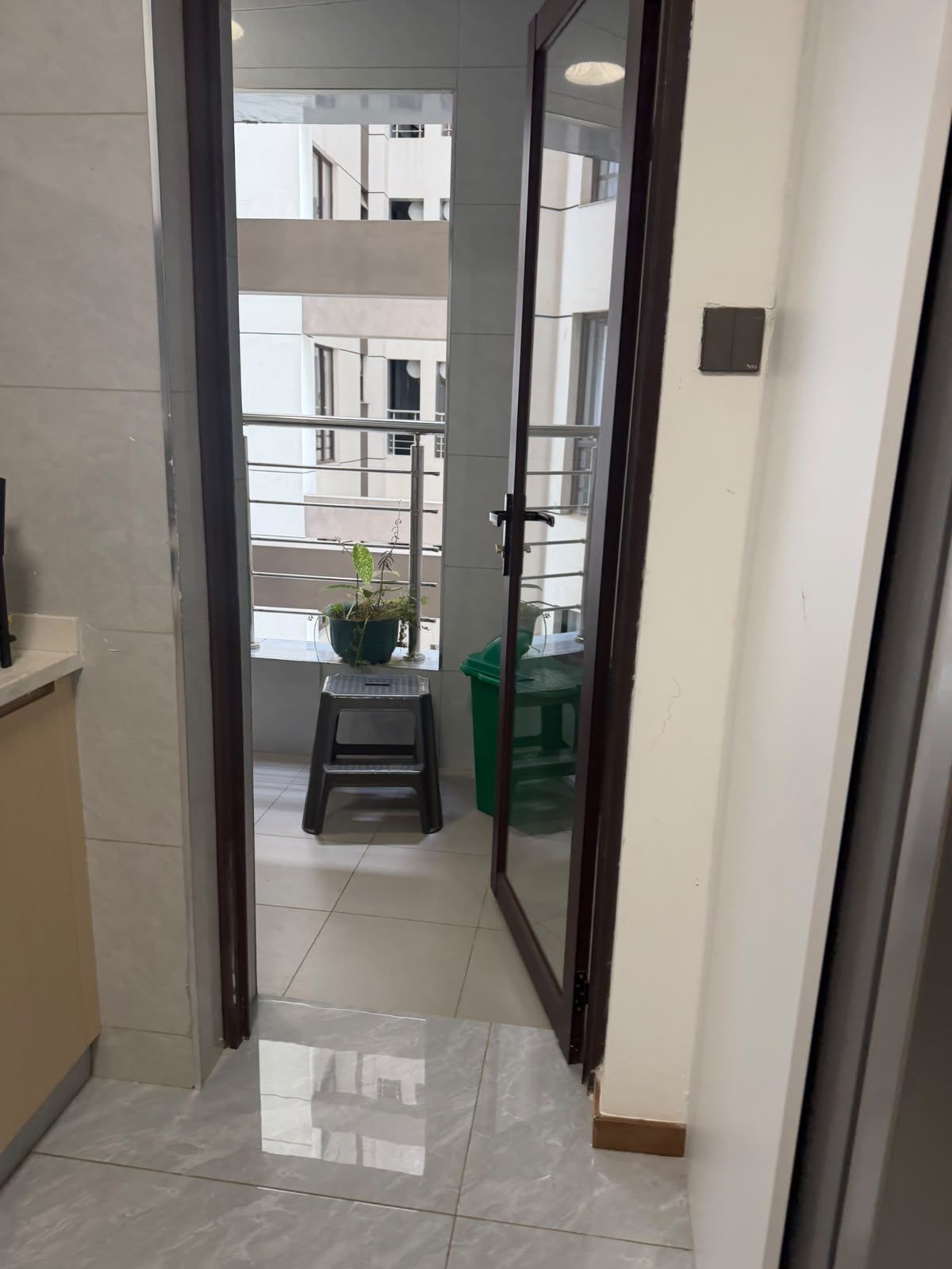A corridor with a tiled floor leading to a balcony with a potted plant and a green trash bin.