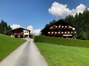 Eine ländliche Straße führt zu einem modernen, zweigeschossigen Haus mit Garage und einem traditionellen Holzchalet mit mehreren Balkonen.