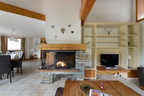 A living room with a stone fireplace, wooden beams, a television, and a dining area with a table set for four. - Vue Montagne | 100 mètres du téléphérique | Sauna (Peisey-Vallandry)