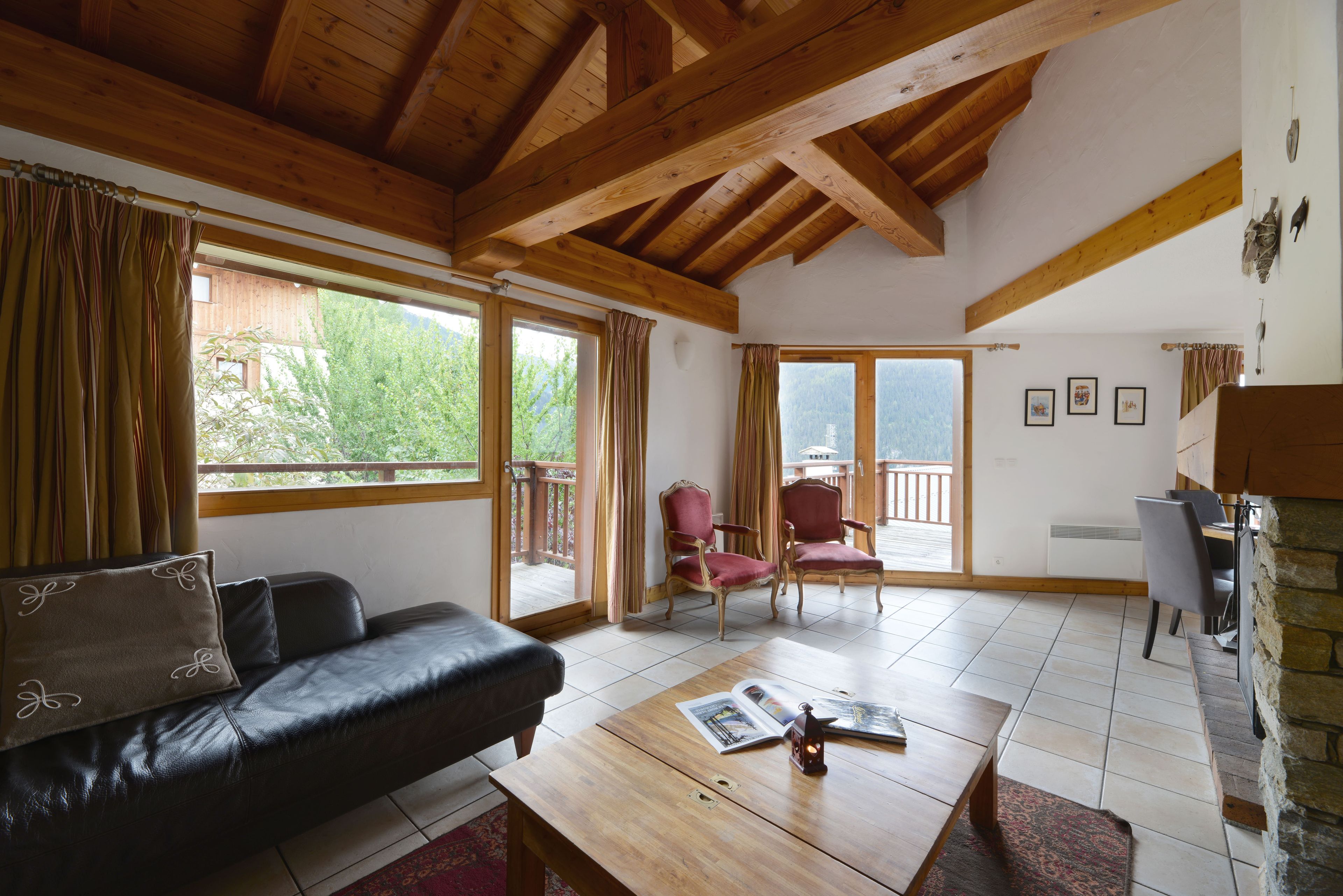 Een kamer met een houten plafond, een zwarte leren bank, een houten salontafel en uitzicht op een balkon met een hekwerk.