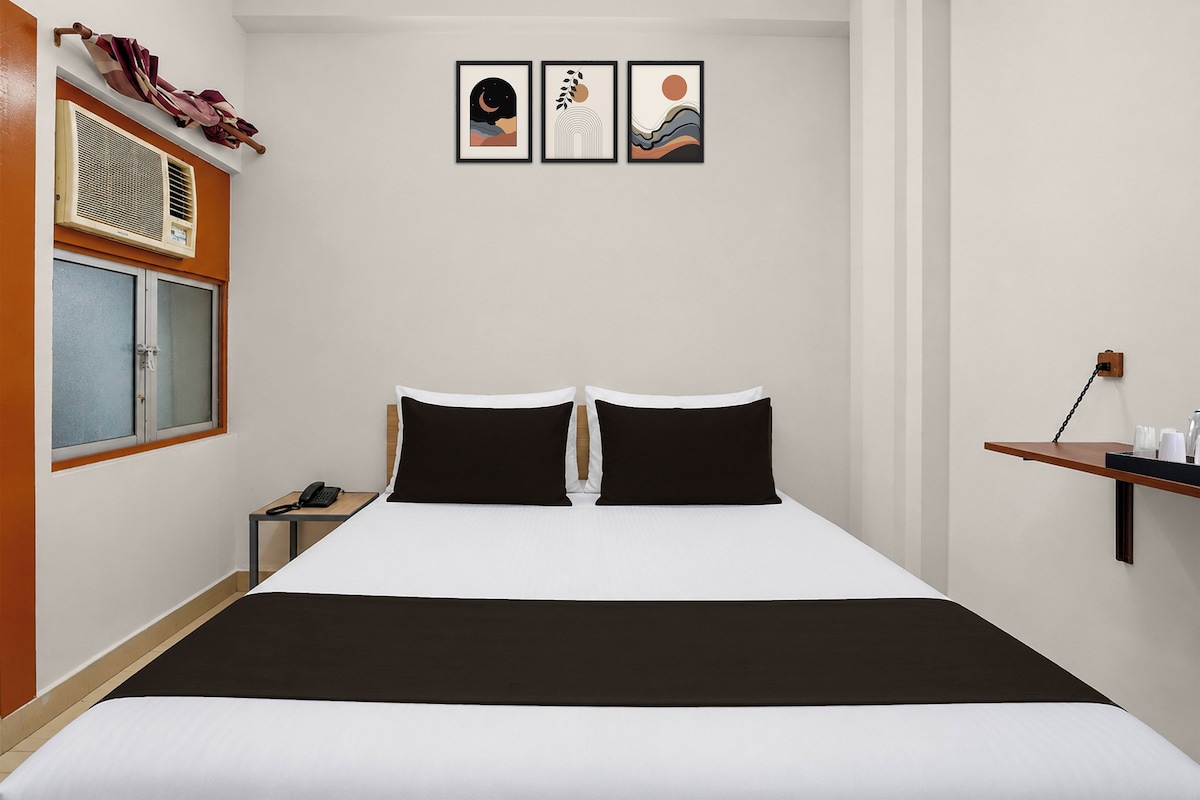 A hotel room with a bed, a nightstand, a telephone, and a window. There are three framed pictures on the wall above the bed.
