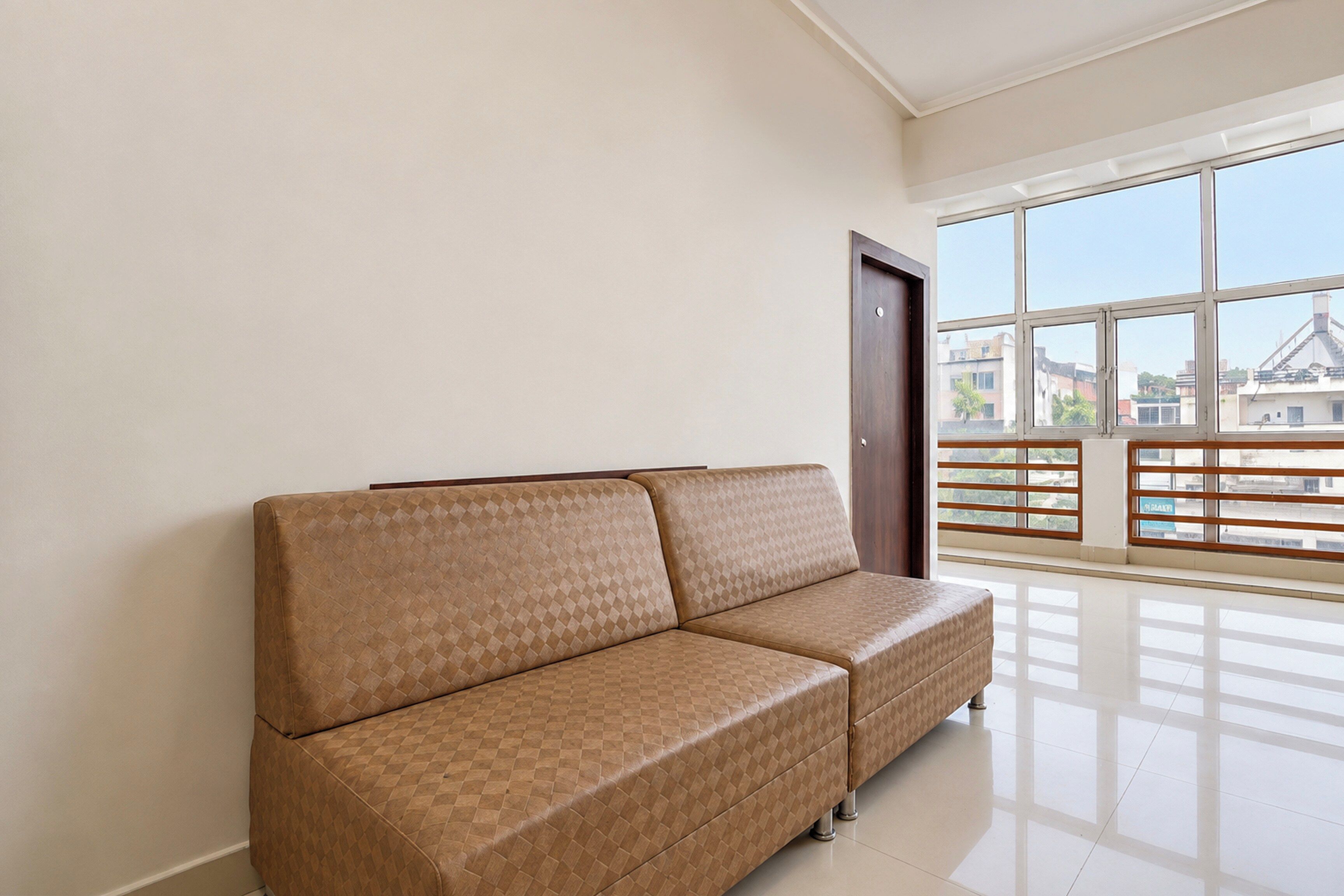 Una habitación con un sofá marrón frente a una pared, ventanas grandes con vista a edificios, y una puerta.
