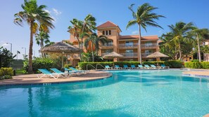Piscina de resort con sillones de descanso y sombrillas, rodeada de palmeras y un edificio de varios pisos.