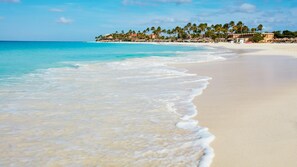 Una playa con agua turquesa cristalina, una orilla de arena y una hilera de edificios con palmeras al fondo.