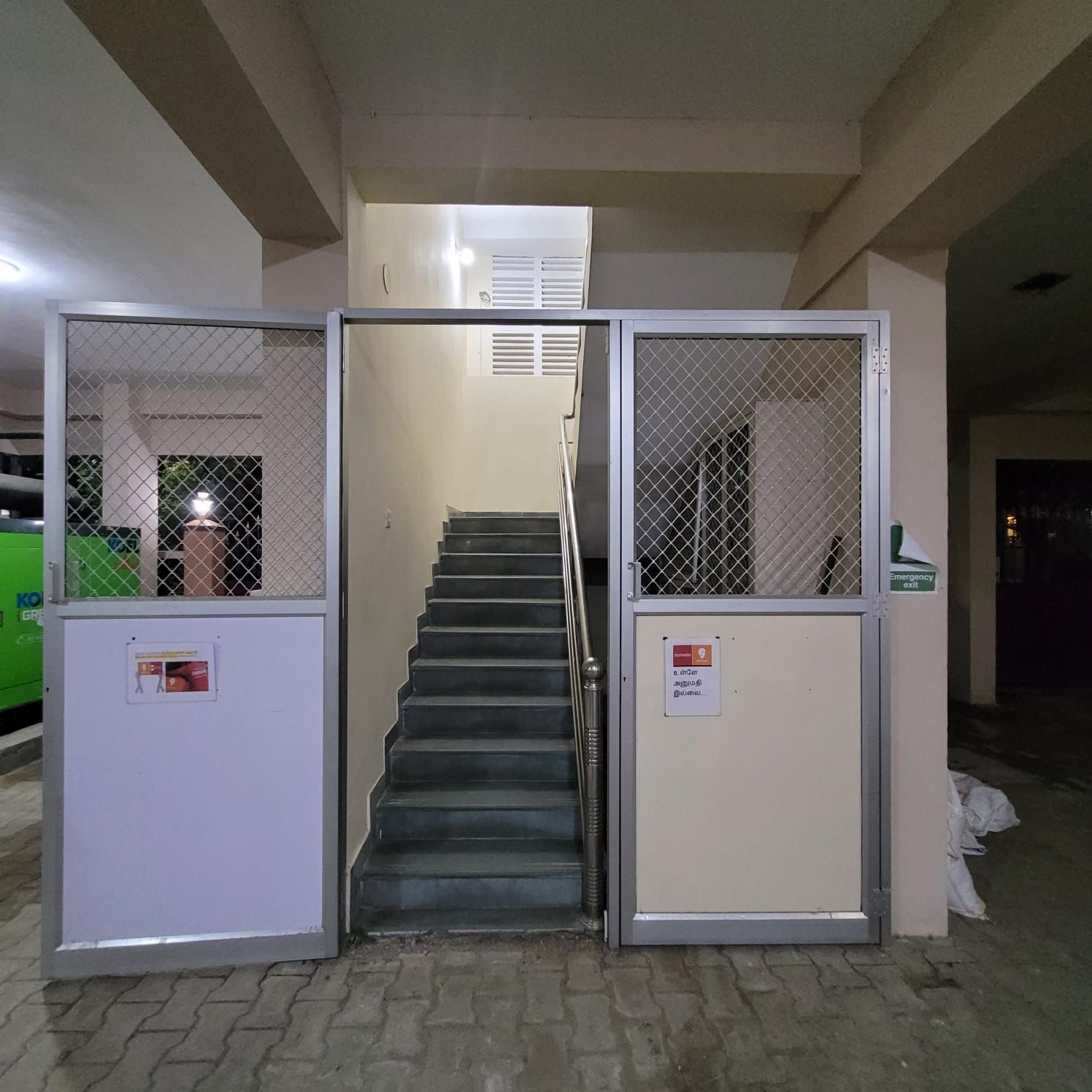 Un escalier menant à un niveau supérieur, flanqué de deux portails métalliques de sécurité portant des panneaux indiquant les sorties de secours.