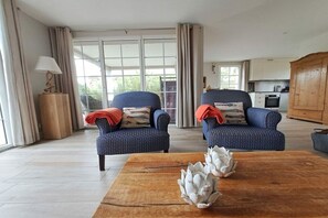 A living room with two patterned armchairs, a wooden coffee table with decorative items, and a view of a kitchen and outdoor area through large windows. - La Petite Dune - A Dream for 2 (Bretteville sur Ay \/ Plage)