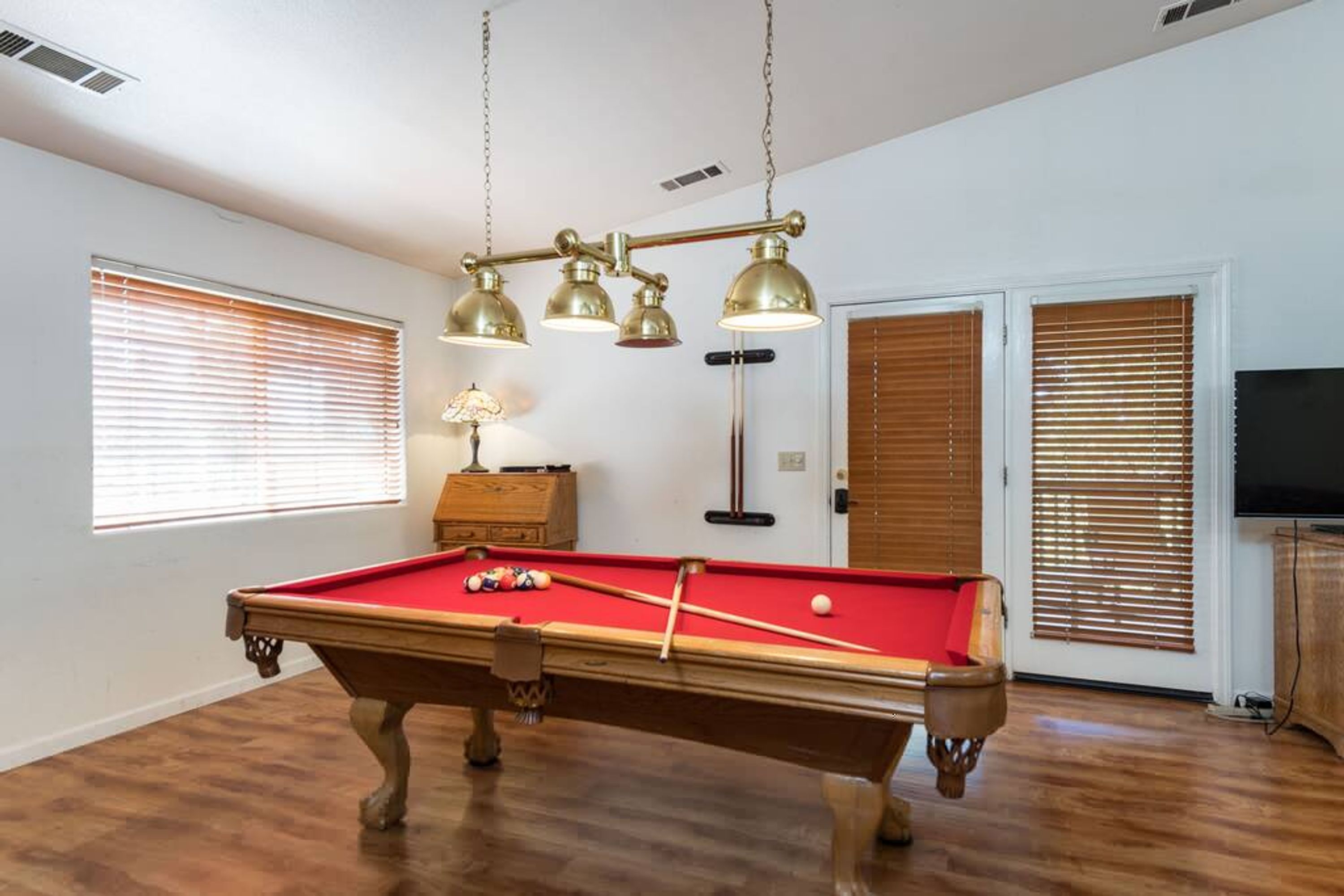 A pool table with balls, a lamp, and a ceiling-mounted light fixture in a room with wooden floors and blinds on the windows.
