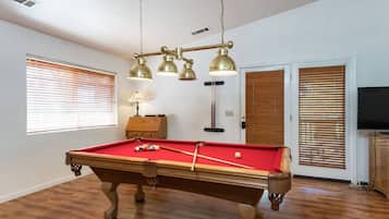 A pool table with balls, a lamp, and a ceiling-mounted light fixture in a room with wooden floors and blinds on the windows.