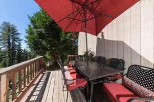 Un patio exterior con terraza de madera, mesa de metal y asientos con cojines rojos.