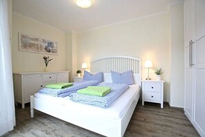 A bedroom with a bed, bedside tables, a nightstand, a lamp, and a wall-mounted artwork. - Beach Villa Marina 18 (Wangerooge)