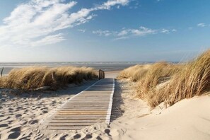 Ein Holzsteg führt zu einem Strand mit Dünen und wolkenlosem Himmel.