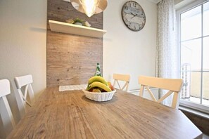 Un comedor con una mesa de madera, sillas blancas, una cesta de frutas, un reloj de pared y un ventanal con cortinas.
