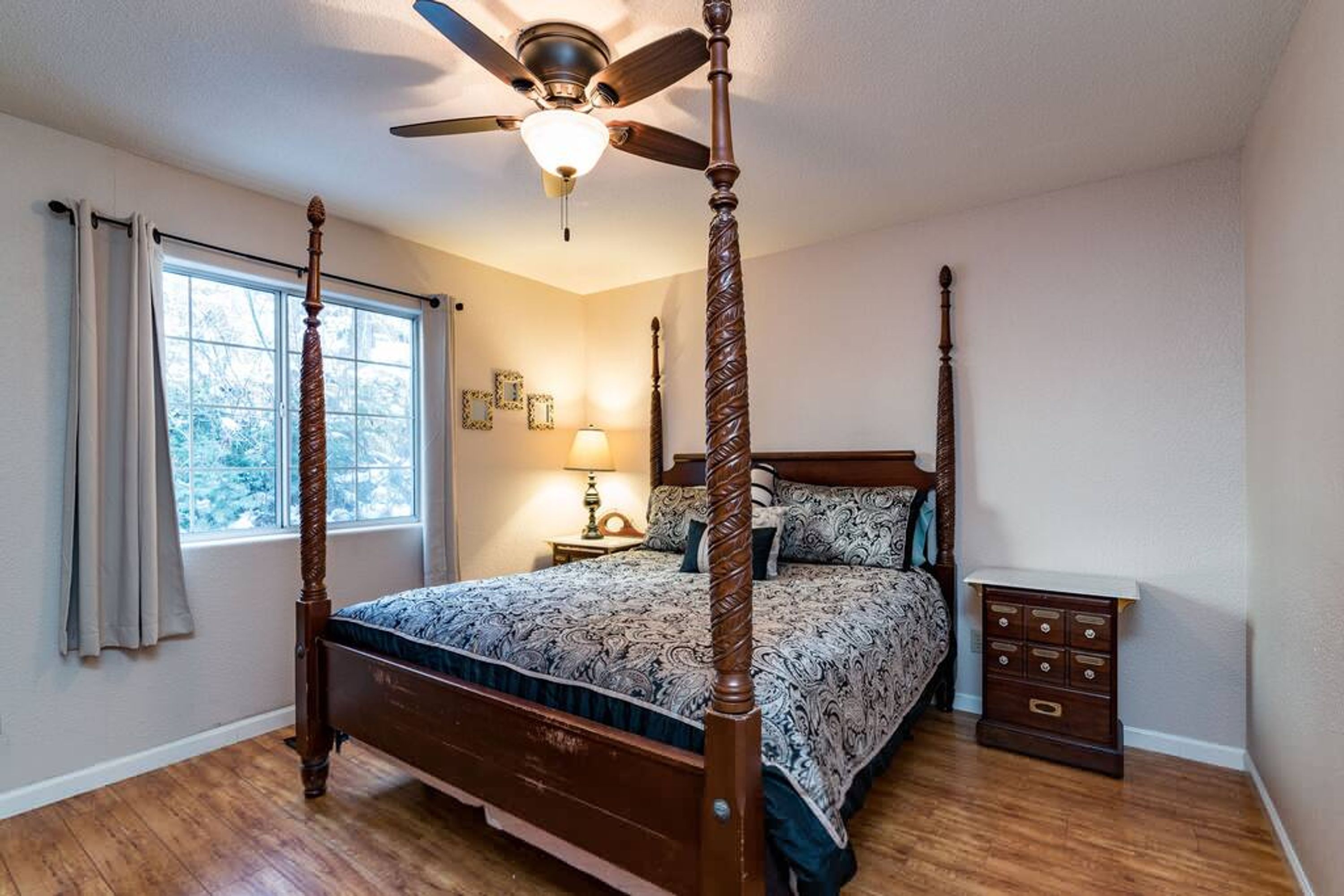 A four-poster bed with a ceiling fan, a bedside table with a lamp, and a dresser with drawers.