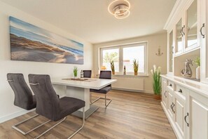 A modern kitchen with a dining area, featuring a white cabinet with glass doors, a painting on the wall, and a view through the window. - View of the dike to the right (Norddeich)
