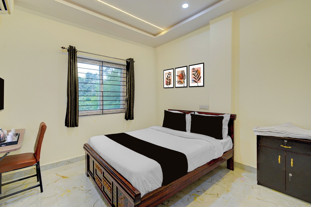 A hotel room with a bed, a desk with a chair, a window with curtains, and framed artwork on the wall.